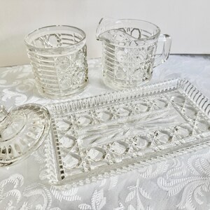 May include: A clear pressed glass set consisting of a lidded sugar bowl, a creamer pitcher, and a rectangular serving tray. Each piece features a repeating geometric pattern of circles and horizontal lines with a starburst center design.
