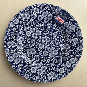Puede incluir: Un plato redondo de cer&aacute;mica con fondo azul oscuro y un denso patr&oacute;n floral blanco. El plato tiene un borde festoneado e incluye una peque&ntilde;a etiqueta rectangular en la esquina superior derecha con el texto Queen's by Churchill Made in England.