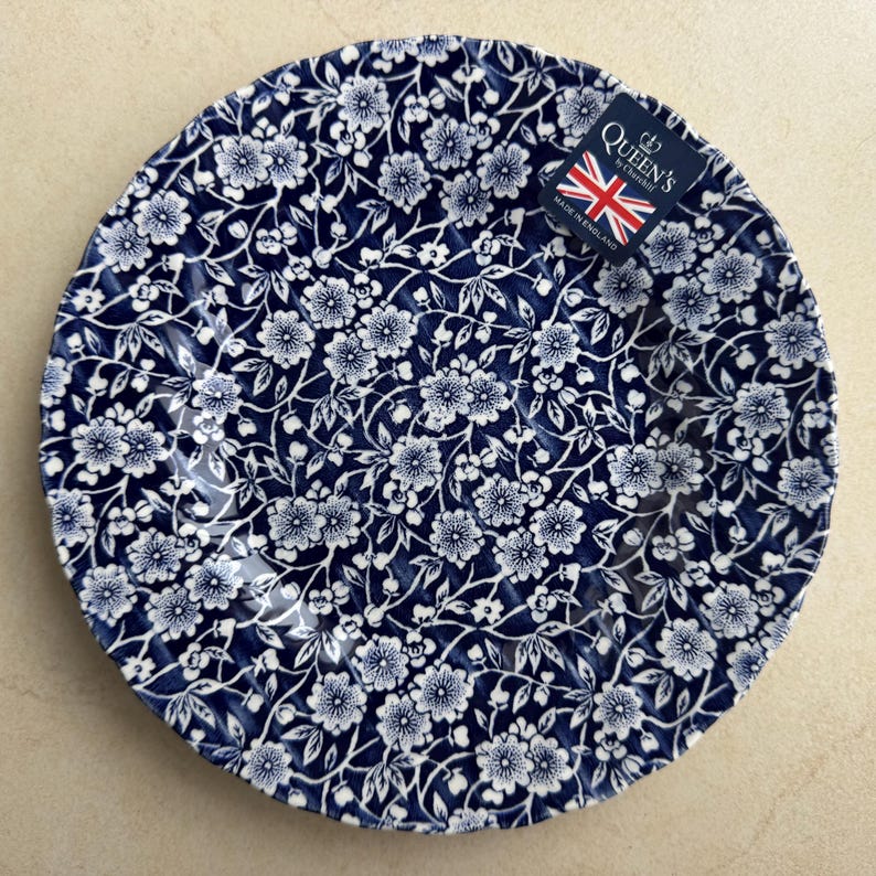 Puede incluir: Un plato redondo de cer&aacute;mica con fondo azul oscuro y un denso patr&oacute;n floral blanco. El plato tiene un borde festoneado e incluye una peque&ntilde;a etiqueta rectangular en la esquina superior derecha con el texto Queen's by Churchill Made in England.