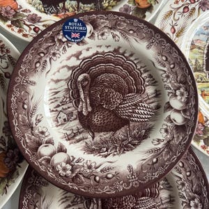 May include: A collection of decorative plates with a turkey design. The plates feature a brown and white colour scheme with a detailed illustration of a turkey in a rural landscape. The plates are made in England.