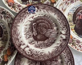 Può includere: Una collezione di piatti decorativi con un motivo a tacchino. I piatti presentano una combinazione di colori marrone e bianco con un'illustrazione dettagliata di un tacchino in un paesaggio rurale. Fatto in Inghilterra.