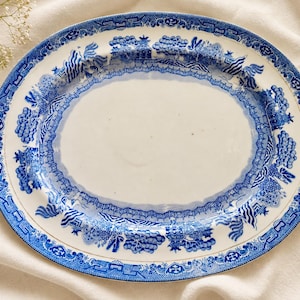 May include: An oval ceramic serving platter featuring a classic blue and white willow pattern. The rim is decorated with intricate illustrations of pagodas, trees, and bridges, while the center of the platter remains a plain, glossy white.