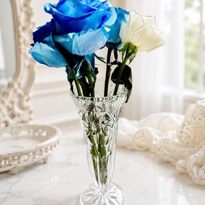 May include: A clear, cut-glass vase with a flared rim and a starburst pattern on the base, containing a bouquet of vibrant blue roses and a single white rose. The glass features vertical fluting and intricate geometric detailing throughout.