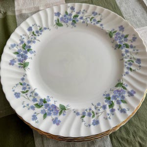 May include: White porcelain plates with a delicate blue floral pattern and gold trim. The plates have a scalloped edge and are stacked on top of each other. The floral design features small blue flowers and green leaves.