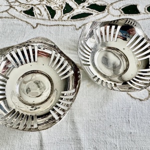 May include: Two silver-toned, decorative bowls with a slotted design. The bowls have a circular base and a wavy, irregular rim. The interior of the bowls reflects light, and they sit on a white surface.