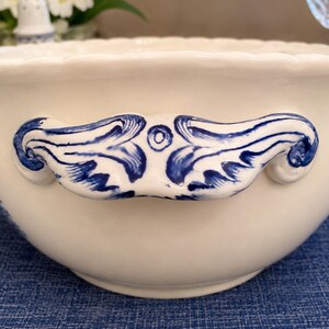 Op de afbeelding: Een cr&egrave;mekleurige keramische kom met een geschulpte rand en twee opvallende handgrepen aan de zijkanten. De handgrepen zijn versierd met een handgeschilderd blauw krulpatroon. De kom heeft een gladde, glanzende afwerking en een ronde vorm.