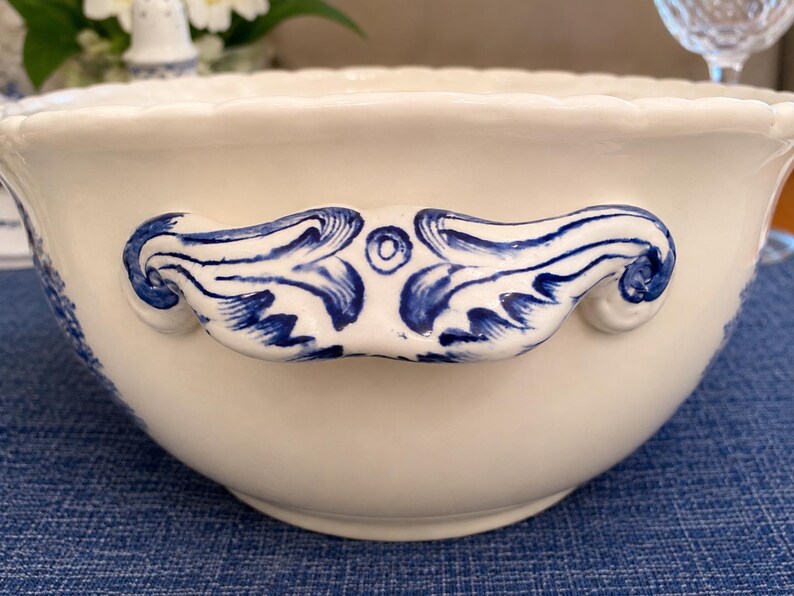 Op de afbeelding: Een cr&egrave;mekleurige keramische kom met een geschulpte rand en twee opvallende handgrepen aan de zijkanten. De handgrepen zijn versierd met een handgeschilderd blauw krulpatroon. De kom heeft een gladde, glanzende afwerking en een ronde vorm.