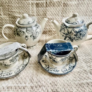 K&ouml;nnte beinhalten: Ein Teeservice mit zwei Teekannen und zwei Tassen- und Untertassen-Sets. Die Teekannen und Tassen haben eine wei&szlig;e Basis mit blauen Blumen- und Vogelmotiven. Die Tassen- und Untertassen-Sets tragen den Text "222 Fifth".