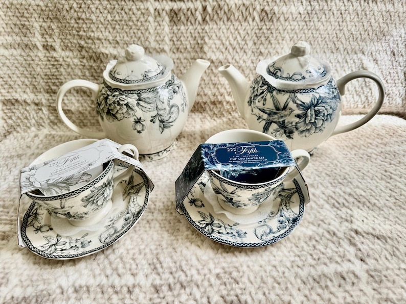 K&ouml;nnte beinhalten: Ein Teeservice mit zwei Teekannen und zwei Tassen- und Untertassen-Sets. Die Teekannen und Tassen haben eine wei&szlig;e Basis mit blauen Blumen- und Vogelmotiven. Die Tassen- und Untertassen-Sets tragen den Text "222 Fifth".