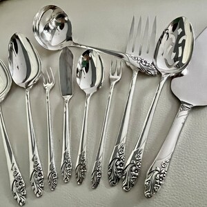 May include: A collection of polished silver serving utensils, including spoons, forks, a knife, and a cake server. The handles feature a floral design. The utensils are arranged on a light gray surface.
