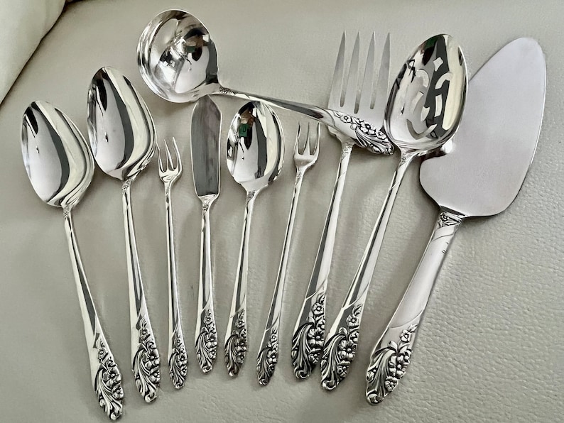 May include: A collection of polished silver serving utensils, including spoons, forks, a knife, and a cake server. The handles feature a floral design. The utensils are arranged on a light gray surface.