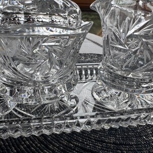 May include: A clear glass set consisting of a footed bowl, a creamer pitcher, and a rectangular tray. Each piece features a cut glass starburst pattern and a scalloped rim. The tray has a textured, diamond-patterned border around its edge.