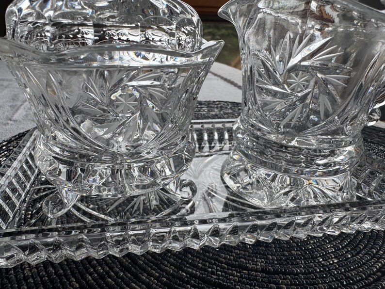 May include: A clear glass set consisting of a footed bowl, a creamer pitcher, and a rectangular tray. Each piece features a cut glass starburst pattern and a scalloped rim. The tray has a textured, diamond-patterned border around its edge.