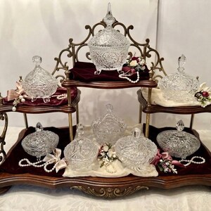 May include: A tiered wooden display stand with ornate metal accents, holding seven clear cut-glass lidded bowls. The bowls feature intricate starburst patterns and are arranged on velvet fabric liners, accented with small floral sprigs and pearl strands.