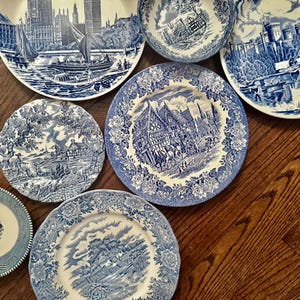 May include: A collection of six ceramic plates and one bowl featuring blue and white transferware patterns. The designs depict various historical landscapes, architectural scenes, and rural settings with intricate floral borders on each piece.