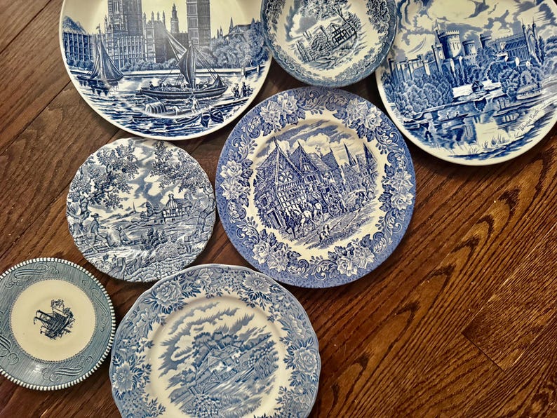 May include: A collection of six ceramic plates and one bowl featuring blue and white transferware patterns. The designs depict various historical landscapes, architectural scenes, and rural settings with intricate floral borders on each piece.