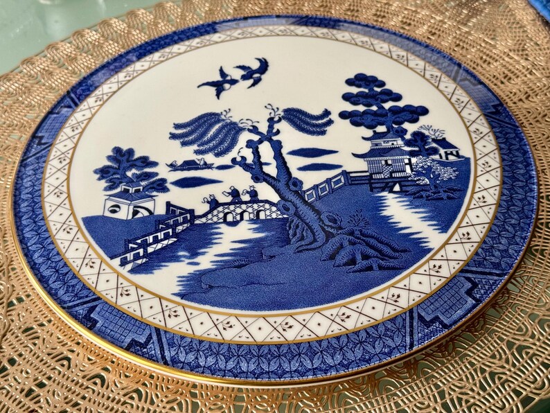 May include: A blue and white porcelain plate with a gold rim and a traditional Chinese design. The design features a bridge, a river, a tree, and a pagoda.