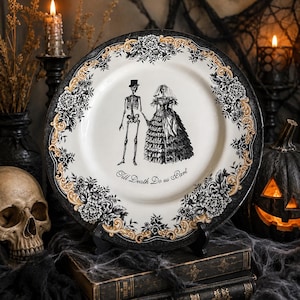 Puede incluir: Un plato redondo de cer&aacute;mica con una ilustraci&oacute;n en blanco y negro de una pareja de esqueletos tomados de la mano, con el texto "Till Death Do us Part" debajo. El borde tiene un dise&ntilde;o floral en negro y dorado sobre fondo blanco.