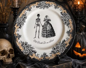 May include: A round ceramic plate featuring a black and white illustration of a skeleton couple holding hands, with the text "Till Death Do us Part" below them. The rim is decorated with a black and gold floral pattern on a white background.