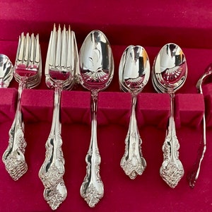 May include: A set of polished silver flatware, including forks, spoons, and serving pieces, arranged in a red velvet-lined case. The silverware features ornate floral detailing on the handles, reflecting light. The set includes various spoon sizes and forks.