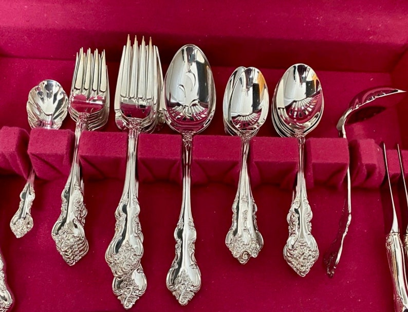 May include: A set of polished silver flatware, including forks, spoons, and serving pieces, arranged in a red velvet-lined case. The silverware features ornate floral detailing on the handles, reflecting light. The set includes various spoon sizes and forks.
