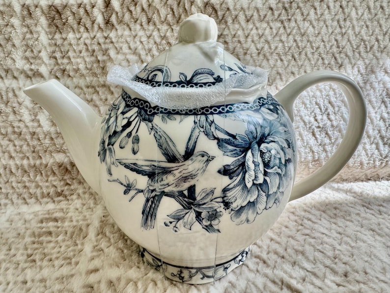 K&ouml;nnte beinhalten: Wei&szlig;e Keramik-Teekanne mit blauem Blumen- und Vogeldesign. Die Teekanne hat einen gebogenen Ausguss, einen abgerundeten K&ouml;rper und einen Griff. Der Deckel ist mit einem kleinen Knopf versehen. Das Design umfasst Blumen und einen Vogel auf einem Ast.