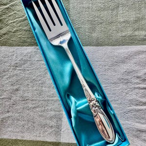 Op de afbeelding: Een zilveren serveervork met sierlijke details op de steel, gepresenteerd in een turquoise satijn gevoerde doos. De vork heeft vier tanden en een decoratief bloemmotief. De doos is rechthoekig en blauw.