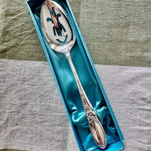 Op de afbeelding: Een zilveren serveerlepel met een decoratief handvat en een gleufkom, gepresenteerd in een blauwe doos met een satijnen voering. De lepel heeft een bloemmotief op het handvat en een uitgesneden patroon op de kom.