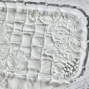May include: A clear glass rectangular serving dish with a woven basket design. The dish features embossed floral and leaf patterns along the edges, including roses, daisies, and grape clusters. The edges have a scalloped design.