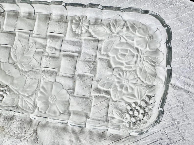 May include: A clear glass rectangular serving dish with a woven basket design. The dish features embossed floral and leaf patterns along the edges, including roses, daisies, and grape clusters. The edges have a scalloped design.