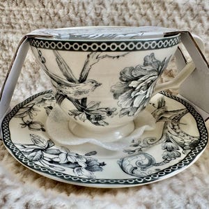 K&ouml;nnte beinhalten: Ein wei&szlig;es Tee-Set mit einem schwarz-wei&szlig;en Vogel- und Blumendesign. Die Teetasse hat einen Henkel und steht auf einer passenden Untertasse. Das Set ist teilweise in einem Karton verpackt.