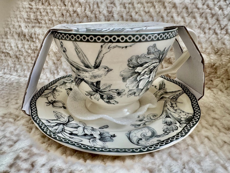 K&ouml;nnte beinhalten: Ein wei&szlig;es Tee-Set mit einem schwarz-wei&szlig;en Vogel- und Blumendesign. Die Teetasse hat einen Henkel und steht auf einer passenden Untertasse. Das Set ist teilweise in einem Karton verpackt.