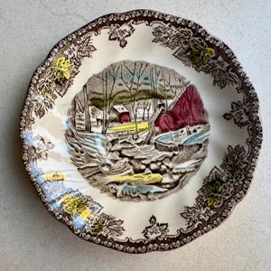 Pode incluir: Um prato redondo de cer&acirc;mica com borda recortada e uma moldura floral castanha. O centro retrata uma paisagem de inverno com &aacute;rvores despidas, uma pequena casa e uma estrutura vermelha, acentuada com toques de tinta amarela, azul e vermelha.
