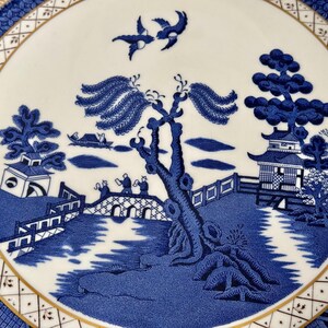 May include: A blue and white porcelain plate with a gold rim features a traditional willow pattern design. The pattern includes a willow tree, a bridge, a pagoda, and birds.