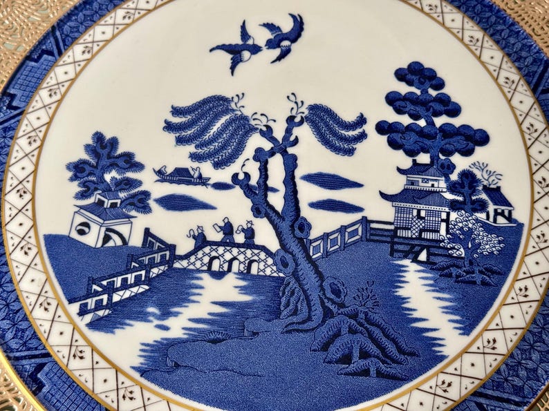May include: A blue and white porcelain plate with a gold rim features a traditional willow pattern design. The pattern includes a willow tree, a bridge, a pagoda, and birds.