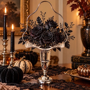 May include: A metallic silver-toned pedestal vase with a scalloped rim and a tall arched handle, filled with a dense arrangement of dark black roses, small black berries, and delicate black foliage, creating a monochromatic floral display.