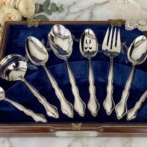 May include: A set of silver serving utensils, including spoons and a fork, displayed in a wooden box with a blue velvet lining. The utensils have ornate handles and are arranged neatly. The box is decorated with butterfly accents.