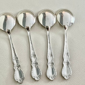 May include: Four silver soup spoons with rounded bowls and ornate, detailed handles. The spoons are arranged in a row, reflecting light. The spoons are likely vintage or antique, and are made of a shiny, reflective metal.