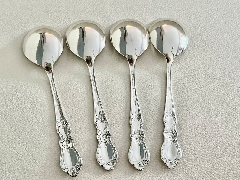 May include: Four silver soup spoons with rounded bowls and ornate, detailed handles. The spoons are arranged in a row, reflecting light. The spoons are likely vintage or antique, and are made of a shiny, reflective metal.