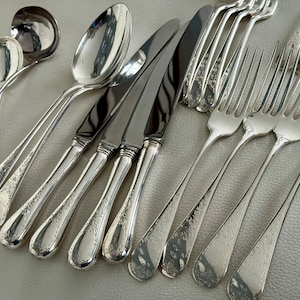 May include: An assortment of antique silverware, including spoons, knives, and forks, arranged on a light-colored surface. The silverware has a shiny, reflective finish and features ornate floral detailing on the handles. The set includes serving spoons, dinner knives, and forks.