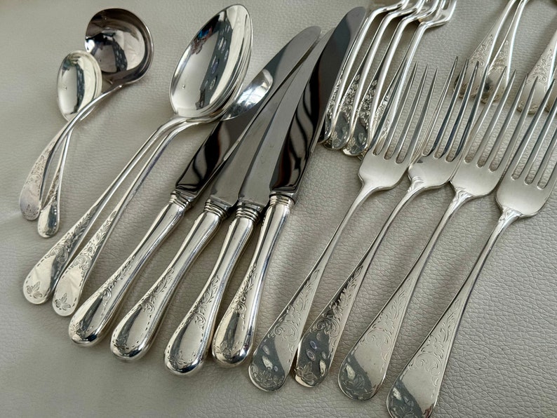 May include: An assortment of antique silverware, including spoons, knives, and forks, arranged on a light-colored surface. The silverware has a shiny, reflective finish and features ornate floral detailing on the handles. The set includes serving spoons, dinner knives, and forks.