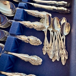 May include: A collection of ornate silver-colored flatware, including spoons and knives, arranged in a blue velvet-lined case. The silverware features detailed floral and scrollwork designs, reflecting a classic aesthetic. The spoons and knives are neatly organized.