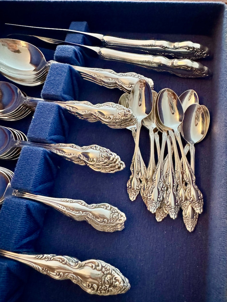May include: A collection of ornate silver-colored flatware, including spoons and knives, arranged in a blue velvet-lined case. The silverware features detailed floral and scrollwork designs, reflecting a classic aesthetic. The spoons and knives are neatly organized.