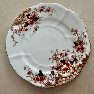 May include: A white ceramic saucer featuring a scalloped edge and a decorative floral pattern in shades of red, brown, and black. The design includes intricate gold-toned accents and small white floral details along the rim of the plate.