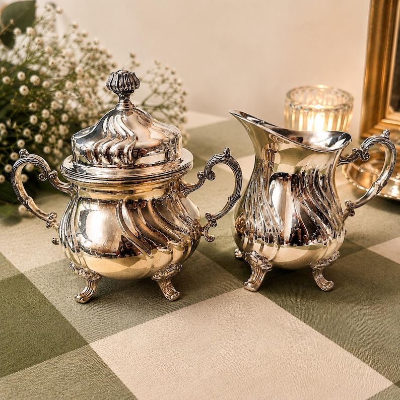 May include: A silver-plated sugar bowl with a lid and a matching creamer pitcher. Both pieces feature a polished metallic finish, ornate curved handles, and decorative feet, with a swirling embossed pattern across the rounded bodies.