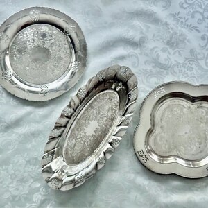 May include: Three antique silver serving trays. The trays have ornate floral and scrollwork designs. One is round, one is oval, and one is a scalloped square shape. The trays are displayed on a white patterned tablecloth.