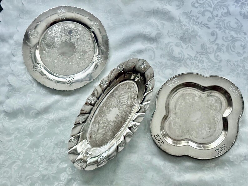 May include: Three antique silver serving trays. The trays have ornate floral and scrollwork designs. One is round, one is oval, and one is a scalloped square shape. The trays are displayed on a white patterned tablecloth.