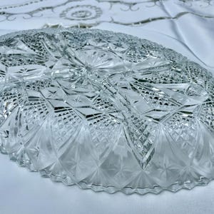 May include: Clear, oval-shaped crystal serving dish with intricate diamond and starburst patterns. The dish has a scalloped edge and is displayed on a white surface. This decorative piece is ideal for serving appetizers or desserts.