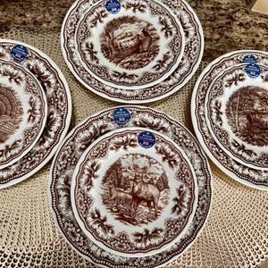 Op de afbeelding: Een set van vier Royal Stafford keramische borden met bruine transferware wildillustraties van een kalkoen, een fazant, een hert en een vogel. Elk bord heeft een geschulpte rand met een bloemmotief en een blauwe sticker.
