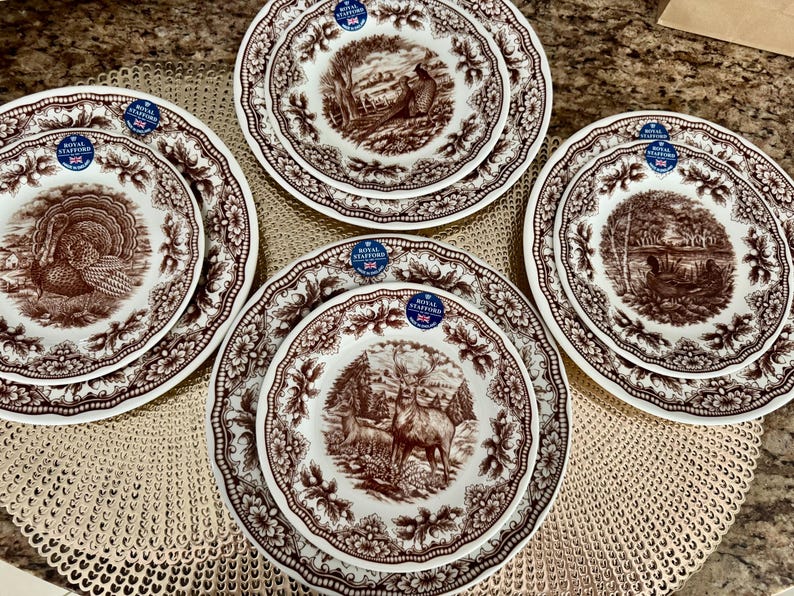 Op de afbeelding: Een set van vier Royal Stafford keramische borden met bruine transferware wildillustraties van een kalkoen, een fazant, een hert en een vogel. Elk bord heeft een geschulpte rand met een bloemmotief en een blauwe sticker.
